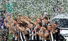 Collingwood players hold the premiership cup on stage with black and white confetti whirling around them