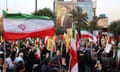 A crowd of people, some holding the Iranian flag. A large image of Ismail Haniyeh is displayed on the side of a building