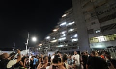 People evacuated from their apartments stand outside the housing complex: it is dark and the multi-storey block with floors arranged in a stepped effect is illuminated against the black sky. Children are seen in adults' arms and people are taking photographs.