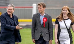 Keir Starmer grins and points at Keir Mather, who wears a Labour rosette on his suit, with Angela Rayner to his right as they all walk on the grass