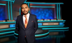 Amol Rajan in a. suit standing in front of a colourful set in blue tones, with two dfour-person desks side by side showing the names of two universities