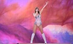 Tall white woman in shimmery white iridescent body suit and knee-high white boots, in front of pink, purple, white and red screen, one arm raised, singing into a microphone held in the other.