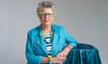 Prue Leith, hands clasped, leaning on turquoise cloth-covered table, against grey background, May 2024