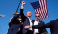 A man with blood running down his fist gestures toward the air with a fist, while secret service agents huddle around him