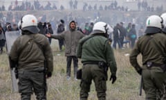 An asylum seeker stands with arms outstretched as police in riot gear stand facing him. Other asylum seekers are in the field behind