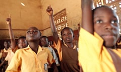 Children dressed in yellow school shirts sit in rows, some with raised hands ready to answer a question.