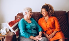 An elderly woman and a young woman sit on a sofa together laughing.