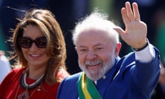 An older bearded man wearing a suit and a green and yellow sash with a younder woman beside him waves to onlookers