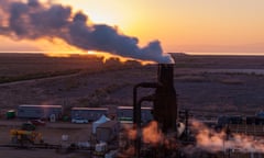 An aerial view of a pink sunset and a smokestack (steamstack?) in silhouette in the foreground, pumping out what is apparently steam.