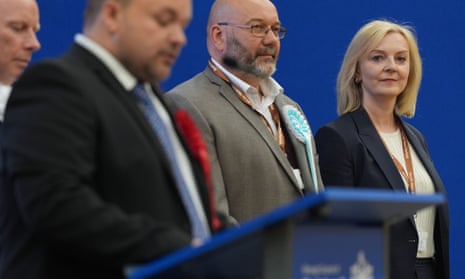Liz Truss, with straight shoulder-length hair and wearing a suit jacket with a ribbon reading "South West Norfolk" smiles tensely as she stands next to two men in suits