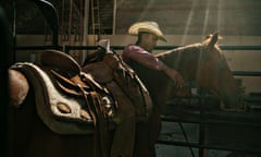 A young Black man wearing a straw-colored cowboy hat lit by sunlight coming through rafters out of the frame has his arm over the neck of a brown, saddled horse.