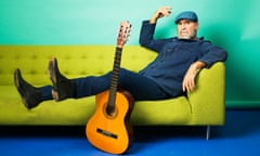 Eric Cantona, with a beard and wearing a beret, stretches out in the corner of a sofa, one arm up and one leg resting on an acoutic guitar