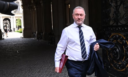 Steve Reed, the secretary of state for environment, food and rural affairs arrives in Downing Street: he is walking through a dark gateway with tall door; a light courtyard is seen behind him and a camera is being pointed at him. He is 60 with short-cropped grey hair and beard, and wears a white shirt and tie; he is carrying his jacket and a red folder