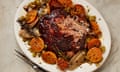 An oval dish of lamb surrounded by sliced tangerines on a marble surface with a fork, knife, glass of white wine, a small plate of tangerine slices and a medium plate of anchovy fillets and shallots