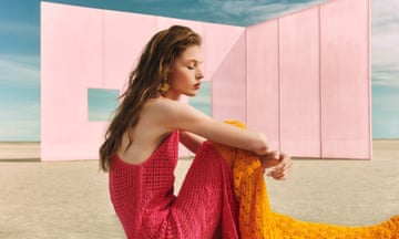 A model wearing a hot pink and orange crocket knit dress sitting down in front of a pale pink wall