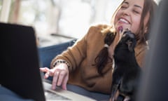 Woman lying on a sofa looking at her laptop, smiling. A small dog licking her face.