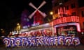 Can-can dancers perform in red, white and blue costumes front of the Moulin Rouge cabaret club at night with illuminated signs and windmill sails