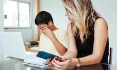 A stock photo in a brightly lit room where a woman who looks white, with long blond hair and holding a cellphone, looks to her left at a middle-schooler who appears Latino, who is covering his eye with one hand and appearing to cry, as he sits in front of a white laptop.