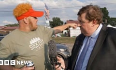 A man wearing a black suit holds up a microphone as he interviews a man wearing a tan T-shirt and a visor as he points to the side