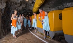 Two men step off a train in a tunnel wearing swimming trunks and holding towels next to others similarly dressed