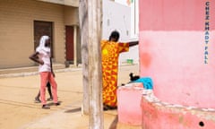 Daylight street scene in Tonghor neighbourhood of Dakar, Senegal. Two men stand with their backs to the camera facing down a dirt street. A pink-washed wall in the foreground.