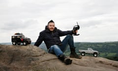 Ian in jeans and hiking boots with two remote-control trucks on top of a rock with valleys behind him