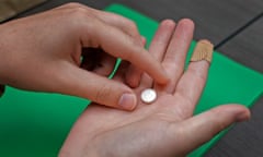 A young white right hand, palm up and a Band-Aid on the index finger, holds a white pill, while the left hand pinches as if to pick it up. Below is a table with a kelly green file folder.