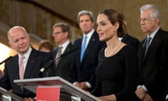 Men in dark suts gather around Angelina Jolie, who is wearing a dark dress and speaking from behind a metal lectern.