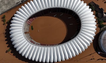 Aerial view of a football stadium where the pitch and all but the highest rows of seats are covered with muddy flood water. The water is also flooding the area outside the stadium,