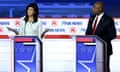 A white woman and a Black man stand at neighboring lecterns on a brightly lit stage. The woman is speaking, with both palms raised, as the man listens.