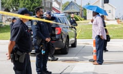 A Black man holding an umbrella, sands in the middle of a street on one side of police yellow tape, speaking toward 3 to 4 uniformed police officers on the other side.