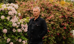 Robert Carlyle standing on a driveway, hands in jacket pockets, flowering bushes behind him