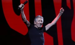 Older white man, short and wiry, wearing black T-shirt and jeans, raises both arms apparently toward crowd, one holding a microphone.