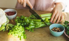 Woman chops a bunch of celery with a knife on a kitchen counter, with a small bowl of nuts and some cauliflower on the side