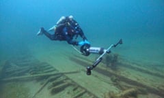 Underwater image, in cloudy blue water, of diver in wetsuit and scuba gear holding a wide pole that seems to have a fan in the middle, perhaps as if it's pulling him across the boards of wood on the seabed below him.