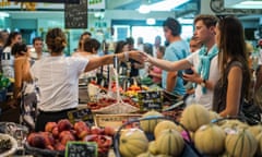 a young male and female purchase food from a vegetable stall in a food market