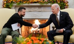 Two white men sit in matching armchairs, leaning forward to shake hands in front of a roaring fire - lot of flowers.