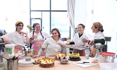 Five young female bakers gather in front of a work table with decorated cakes.