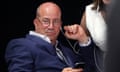 Jeff Zucker listens to a presidential debate hosted by CNN in 2019: he is holding a phone and has an earpiece in one ear; the other earpiece is worn by a woman who is partially seen in the background leaning over him. He is sitting in a relaxed posture and wears a navy blue jacket and open-necked white shirt; he is a middle-aged man with bald head and wears circular glasses.