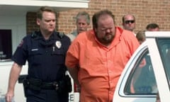 Man in orange jumpsuit is escorted to white police car by police officer wearing dark uniform, belt and gun