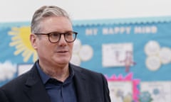 A man wearing a suit and glasses stands in a primary school classroom in front of a bright blue display of artwork