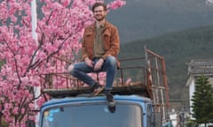 Alec Ash sitting on top of his blue, beaten up van, in rural China, the Cangshan mountain behind him, a cherry blossom tree in full bloom to the side.