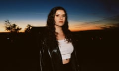 Bethany Cosentino wearing a white top and black leather jacket, standing outside at dusk, the evening sky dark blue and orange, trees in silhouette.