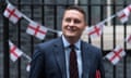 Wes Streeting leaves Downing Street after attending a cabinet meeting: he is seen in a head and shoulders view in front of black iron railings on which England flags are hung as bunting. He is a young man with short fair hair and is smiling; he wears a dark blue jacket, pale blue shirt and dark tie with red horizontal stripes, and holds a red folder under his arm.