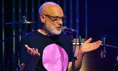 Brian Eno at the Royal Festival Hall, London, wearing a black t-shirt with a big pink circle on it, talking into a microphone with his hands upturned