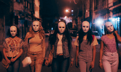 six young women side by side in identical white carnival masks walking towards the camera, down a street at night