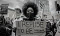 A young black protester with a big 'afro' hairstyle and a black bandana over her face holds a cardboard sign to the camera reading "I refuse to be silent"