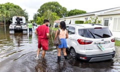 Two people stand in ankle-deep water next to a car on a flooded residential street, as a man further up the street stands next to a recovery truck