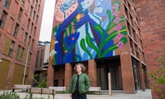 Bryony Bond, in a puffer jacket and with short curly hair, stands in front of a brick building with a mural of swirly plants on it