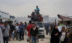 Crowds of people walk along a busy street. A small van is being driven with belonging strapped to the roof and people sitting on top of it